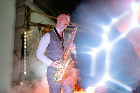 A formally dressed musician plays saxophone on a dark industrial stage, surrounded by dramatic spotlights and swirling smoke