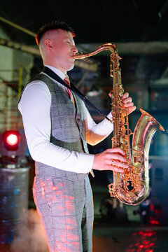 Caucasian man professional musician performs a jazz saxophone solo in a smoky, dark industrial venue, bathed in a single bright spotlight while wearing formal attire
