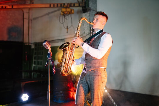 Solo musician in a formal vest and dress shirt plays a golden saxophone under glowing stage lights in an urban industrial environment while entertaining an audience at a dark live music event venue