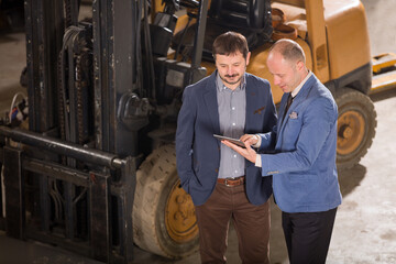 High angle view of two businessmen without hard hats standing in front of a forklift and looking at a digital tablet inside a warehouse. © Fatih