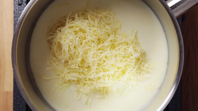 Grated cheese drops into white sauce in saucepan. Cheese sauce works great as base for ham casserole, mac and cheese or vegetable gratin. Cheese sauce brings warm, comforting flavor to any dish.