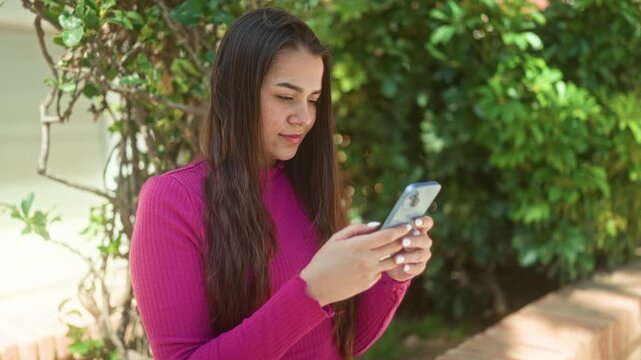 Woman holding smartphone, fingers tapping screen while standing by a brick ledge and leafy hedge near building; digital connection calm.