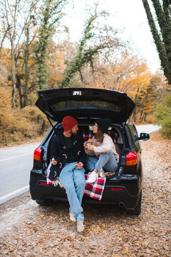 Romantic couple woman and man sitting in car trunk having picnic over nature autumn background. Fall season.