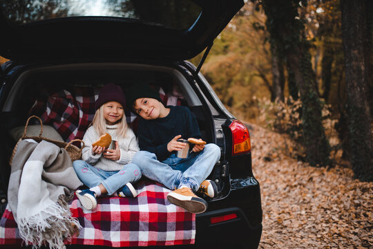 Cute children having picnic in car trunk sitting on checkered blanket in woods over autumn leaves. Fall season. Childhood.