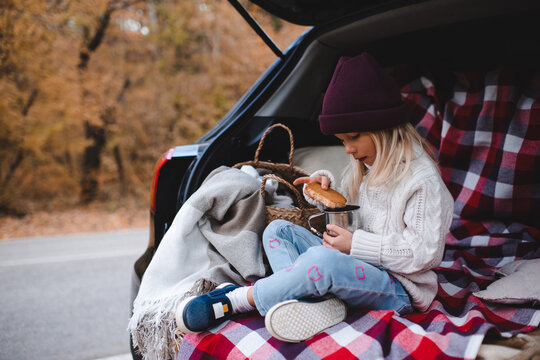Cute child girl 4-5 year old eating biscuits in car trunk sitting on checkered blanket in woods over autumn leaves. Fall season. Childhood.