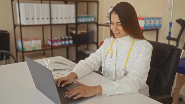 Woman doctor typing on laptop at desk in building, stethoscope around neck with glasses and telephone on table; focused care.