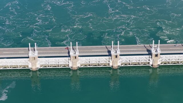 Aerial drone view of a massive storm surge barrier in the Netherlands. Powerful water stream flowing through the opened dam gates in an industrial coastal landscape.