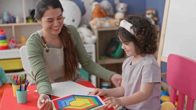 Teacher guiding girl holding colorful wooden puzzle on a red table with colored pencils and stuffed toys in a playroom inside a building; learning joy.