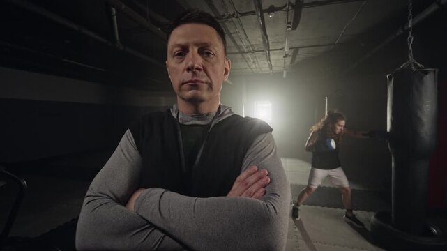 Portrait of Caucasian middle aged male coach standing with crossed arms in gym while Caucasian young woman hits punching bag. Ideal for promoting fitness and personal training services
