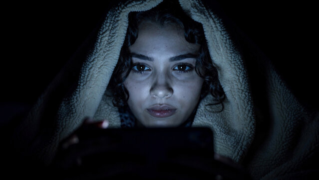 Young woman engrossed in smartphone screen, face illuminated by device light, concept for technology addiction, online engagement and isolation