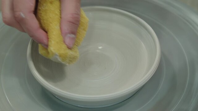 Potter smoothes edges of plate and absorption of excess water of product during work on circle with synthetic sponge. Final stage of work on ceramic product. Handicraft workshop with potters wheel
