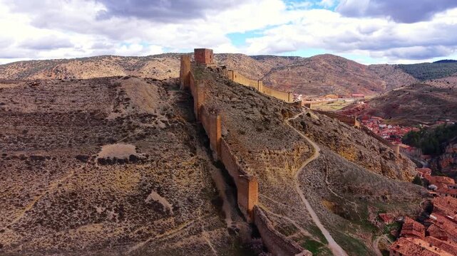 Ancient medieval defensive walls and tower of Albarracin stretching across a rocky hillside in Aragon, Spain, with the historic red-roofed village visible below