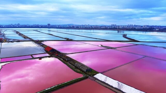 Pink salt pans stretch toward city skyline Bulgaria. Aerial view reveals geometric pools, white salt mounds and cloudy sky.