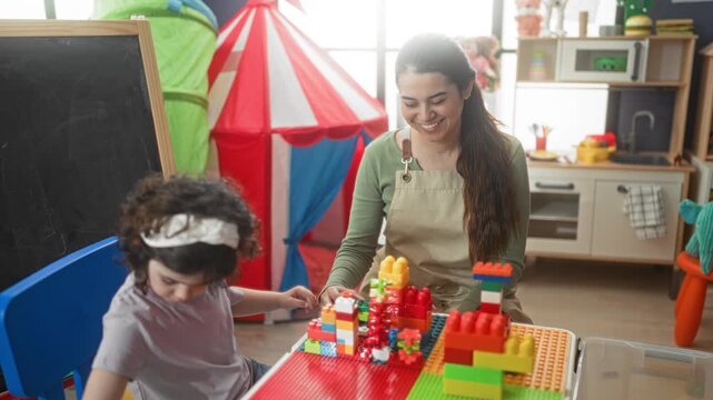 Woman builds colorful towers with large plastic blocks at a table while guiding a child in a preschool classroom with tent and chalkboard visible; joyful learning.