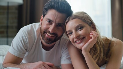 Fototapeta premium Happy Caucasian couple in love enjoying good morning smiling looking at camera cuddling lying in bed. Loving husband boyfriend man with wife woman girlfriend family smile cuddle relaxing in bedroom