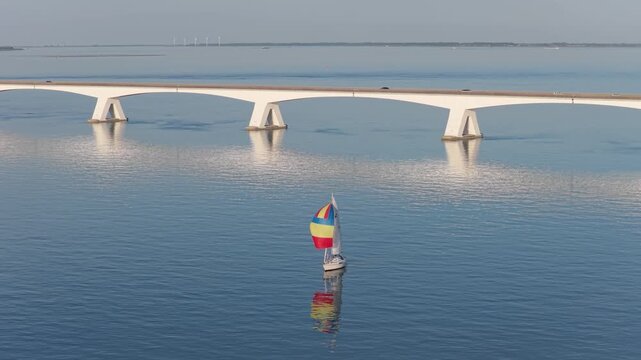 Aerial drone view of a sailboat near a long bridge. Summer vacation, sailing travel, and coastal journey in scenic waters.