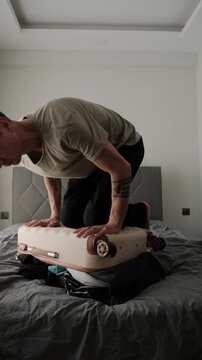 Last minute packing before travel. Man struggling to close overfilled suitcase while preparing for vacation, relocation or business trip, vertical