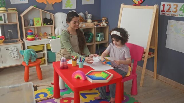 Woman hands paper to girl while child cuts with scissors at a classroom table with easel, toys, crayons and rainbow puzzle; nurturing concentration.