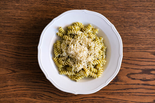 A serving of fusilli pasta with vibrant pesto sauce and a generous heap of grated Parmesan cheese, presented in a white plate on a wooden background. 