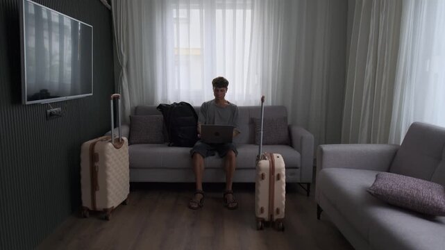 Always working lifestyle and nonstop mobility. Man working on laptop on sofa with luggage, finishing work and leaving apartment immediately