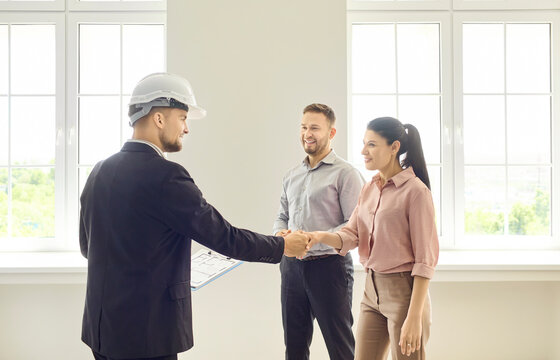 Couple and realtor handshake in apartment consultation. Smiling clients meet agent in bright empty room after inspection, discussing purchase details. Real estate purchase agreement concept.