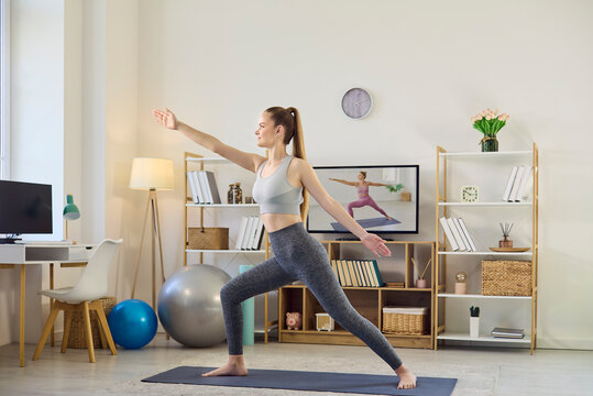 Woman doing yoga home fitness workout online. She follows a trainer on tv in a living room. Modern decor with mat, balance ball, and shelves. Concept of mindfulness, wellness, and wellbeing