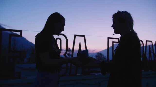 Silhouettes of girls on background of text in popular tourist destination. Love story girl gives gift to girl on background of setting sun