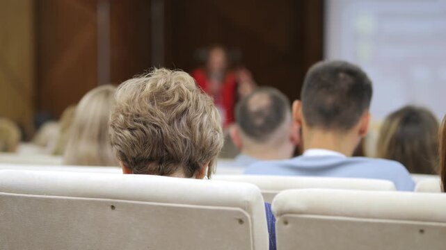 Audience members watch presentation from behind. People focus on speaker, showing interest. Scene shows conference or seminar setting. Ideal for business, education, or corporate events