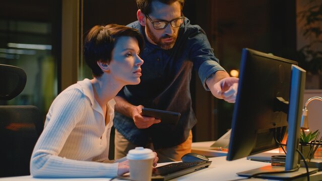 Business team employers with computer discuss work at night evening office Caucasian man leader checking working process help with difficult task woman intern talk with boss pointing at screen monitor
