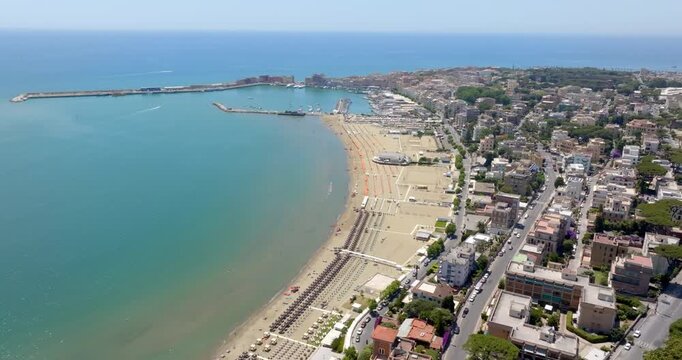 Aerial view of Anzio, Lazio, Italy. It's a coastal city featuring a curved sandy beach, turquoise sea waters, a busy harbor, and urban streets, perfect for a summer travel destination. Sunny morning.