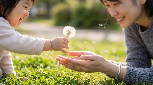 緑豊かな芝生に寝転がりタンポポの綿毛で遊ぶ笑顔の母親と子供