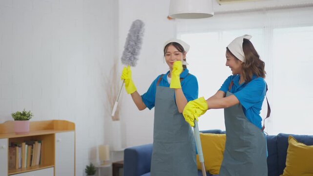 Woman cleaner with duster mop glove apron in living room, cheerful cleaning housework scene, smiling cleaning service team in bright home interior with sofa, window, plant, shelf, soft daylight