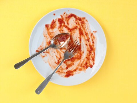 Top view of an empty dirty white ceramic plate with a spoon and fork smeared with red tomato sauce or ketchup against yellow background. Concept of finished meal, food leftovers, and dirty cutlery