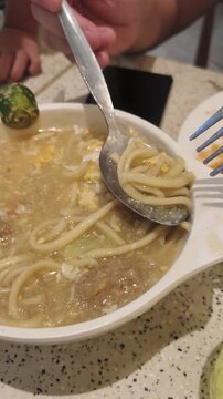 Person lifts spoonful of noodle soup with meatball using fork and spoon