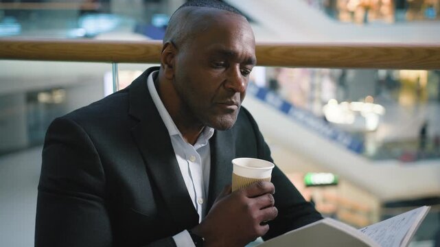 Relaxed mature African American business man drinking coffee reading book in cafe mature calm 50s businessman male visitor client drink tea cappuccino read literature novel in cafeteria break pause