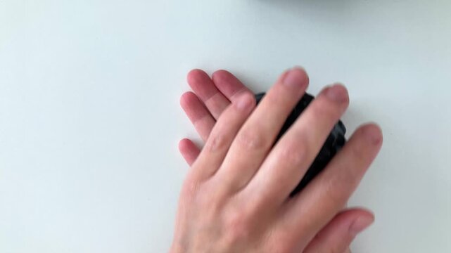 A woman rolls a black massage ball over her palm to relieve tension, warm up her joints, do hand exercises, and relax. Top view of a woman's hands on a white background
