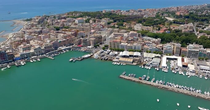 Aerial view of Anzio, in Lazio, Italy. It's a coastal city featuring a docked boats, green-turquoise waters, coastal piers, and dense urban buildings under a clear summer sky, ideal for tourism.