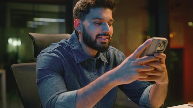 Happy Indian business man browsing mobile phone in evening office at table joyful Arabian businessman smiling scrolling web app Hindu male guy professional smile looking in smartphone at night agency