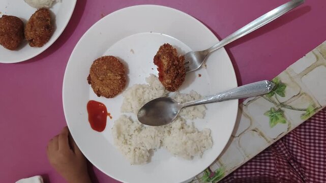 Child uses fork and spoon to eat rice and fried patty with ketchup on plate