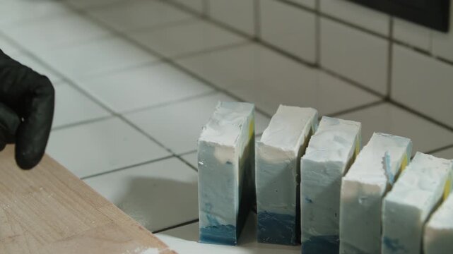 Cutting bar of blue soap into small pieces for packaging and sale. Wooden cutting board, worker's hands in black protective gloves