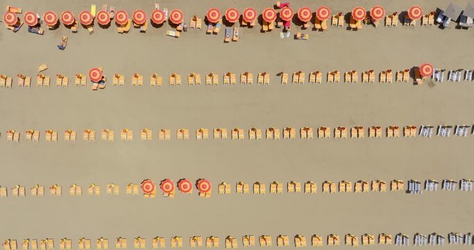 Top-down aerial view showing neat parallel rows of orange umbrellas and sunbeds lined up on golden sand, capturing an organized pattern of summer relaxation at a coastal beach resort. Zenithal shot.