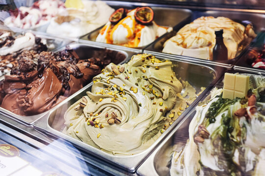 Assorted artisan gelato in display case background. Pistachio chocolate and creamy frozen desserts inside metal trays. Colorful ice cream selection for sweet shop counter. Italian style cold treats.