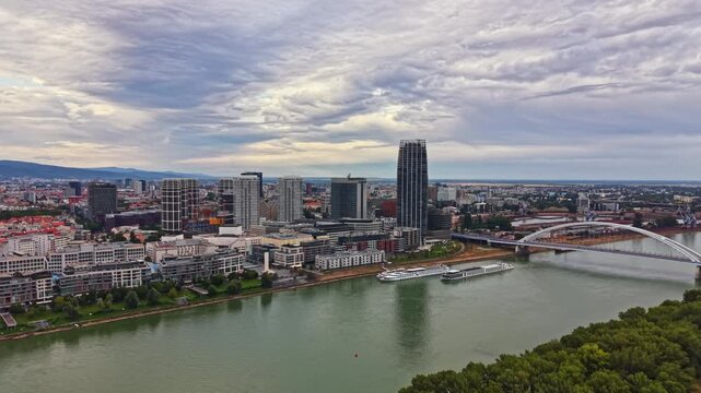 City skyline features tall buildings near a river with a bridge connecting both sides. Clouds fill the sky. Boats are visible floating on the water.