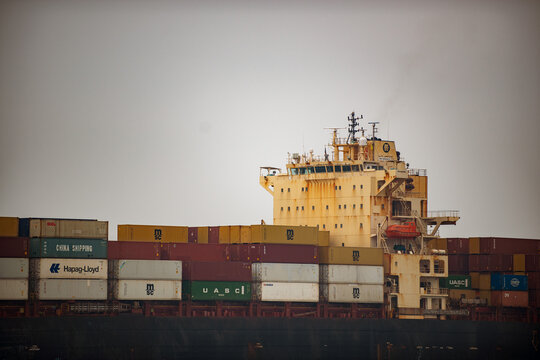 Close-up of the upper deck and stacked cargo containers of a container ship, showing containers from multiple shipping companies (MSC, China Shipping, Hapag-Lloyd, UASC, COSCO) under an overcast sky.