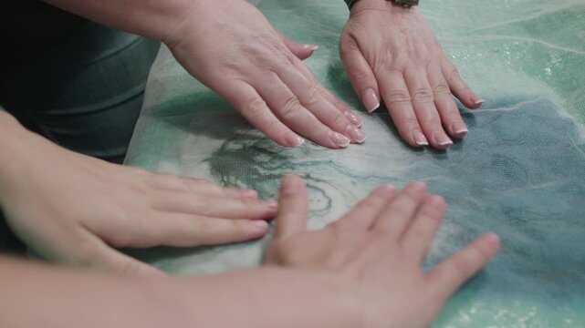 Womens hands press pieces of wool that are between gauze and antistatic film to give density. Master shows student correct movements for working with wool
