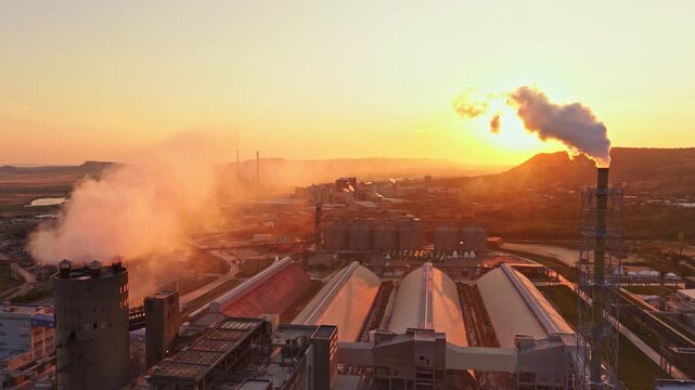 Smoke rises from chimneys as the sun sets behind a large factory complex. The landscape includes silos and long buildings, with distant hills visible.