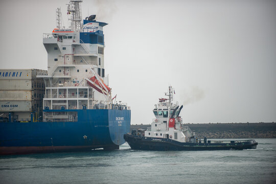 Tugboat "V.B. Deauville" maneuvering alongside the stern of the container ship "CMA CGM White," with visible CMA CGM and Marfret branding on the containers and hull, under a gray sky, in Le Havre