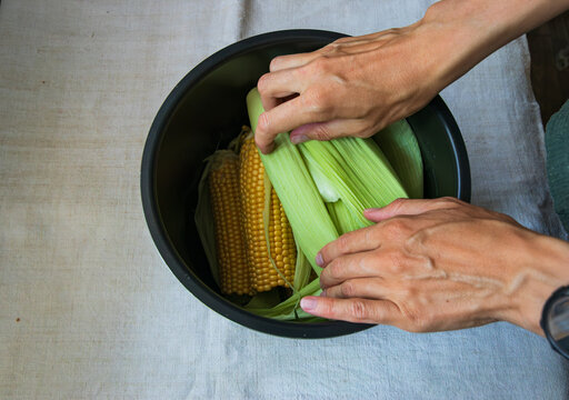 Fresh sweetcorn on the cobs are in a pot. Female hands cover corn cobs with green corn leaves. Sweetcorn cooking. 
