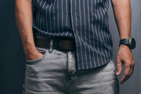 Man wearing grey jeans, leather belt, striped dark shirt and watch with hand in pocket.
