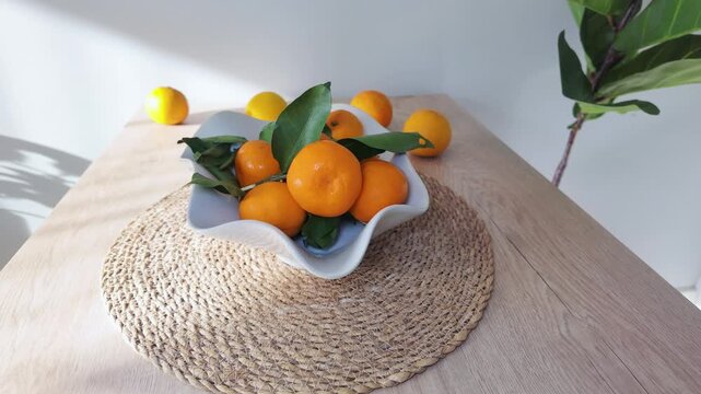 Fresh ripe oranges with green leaves rest in a ceramic bowl on a wooden table.